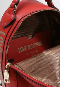 Červená kabelka Love Moschino se zlatým zipem, otevřená a odhalující vzorovaný saténový interiér a značkovou červenou vnitřní kapsu.