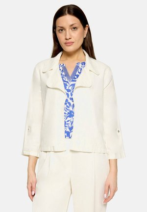Femme aux cheveux bruns raides portant une veste blanche et un pantalon blancs par-dessus un haut à motif floral bleu et blanc, debout devant un fond uni.