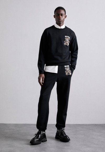 Set de sweat și pantaloni negri cu grafică de urs. Guler rotund, manșete ribbed și accente cu logo. Stilizat cu o cămașă albă cu guler dedesubt.