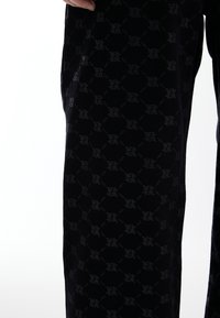 Tissu tricoté noir avec un motif de logo répétitif subtil en gris. Un gros plan montre la texture et la coupe ajustée, sans aucun matériel visible.