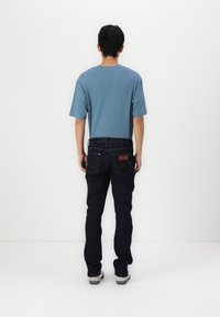 T-shirt de algodão azul com mangas curtas combinada com jeans azul escuro. Os jeans apresentam um remendo de couro castanho e dois bolsos traseiros com detalhes de costura.