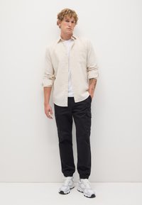 Camisa de botón en color beige claro sobre una camiseta blanca, combinada con pantalones cargo negros y zapatillas blancas. Tela suave y ajuste relajado.