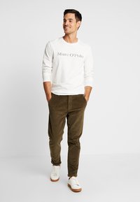 Vit långärmad skjorta med "Marc O'Polo"-logotyp, olivgröna kordbyxor och vita sneakers med bruna sulor, stående mot en grå bakgrund.