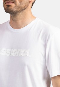 Biała bawełniana koszulka z okrągłym dekoltem, z wypukłym białym napisem "ASSIGNOL" na piersi. Krótkie rękawy i gładka faktura.