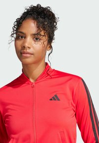 Rode sportieve zip-up jack met zwarte strepen langs de mouwen en een klein zwart logo. Soepele stofstructuur en een getailleerd ontwerp.