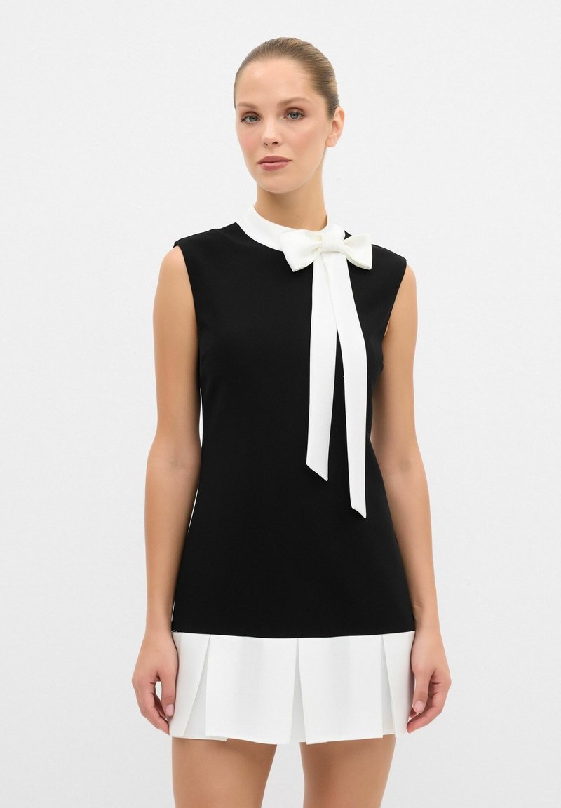 Schwarzes Kleid mit einem weißen Kragen und einer Schleifen-Details, ausgestattet mit einem plissierten weißen Rock. Glattes Material, ärmelloses Design und taillierter Schnitt.