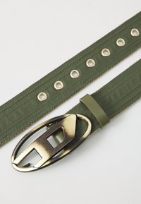 Ceinture en tissu vert olive avec boucle en métal présentant une découpe géométrique. Des œillets espacés le long de la ceinture ajoutent du détail et de la fonctionnalité.