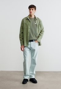 Νεαρός άνδρας που στέκεται μέσα σε κλειστό χώρο, φοράει πράσινο πουκάμισο καρώ πάνω από ένα ελαιί πουλόβερ, ανοιχτό μπλε τζιν, μαύρη ζώνη και μαύρα παπούτσια.