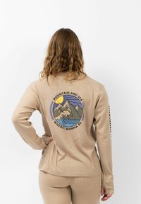 Beige langemouwtop gemaakt van textuurstof, met een geprinte grafiek van bergen, zon en water op de achterkant, met een ronde vorm.