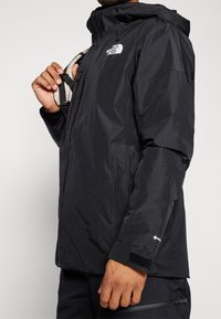 Giacca nera impermeabile con zip frontale, cappuccio regolabile e polsini in velcro. Presenta un logo sul petto e un tessuto testurizzato.