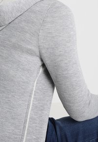 Närbild av en person som bär en ljusgrå långärmad hoodie och mörkblå jeans, sittande med ryggen delvis vänd.