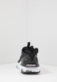 Zwarte sport sneaker met een textiel bovenwerk, glanzende zwarte accenten, een trekstrip en een chunky witte zool met grijze details aan de onderkant.