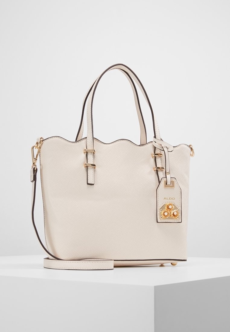Beige textured handbag with scalloped top, gold hardware, detachable strap, and decorative charm featuring details in gold and orange.