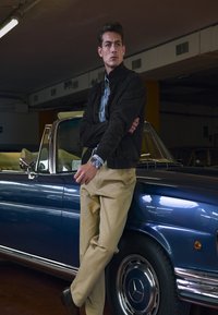 Man with crossed arms leaning against a blue vintage Mercedes convertible in a dimly lit indoor parking area.