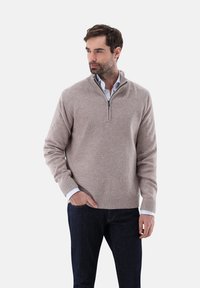 Beiger Pullover mit halbem Reißverschluss, gerippten Bündchen und Saum, aus weich gestricktem Stoff, getragen über einem Kragenhemd und dunklen Jeans.