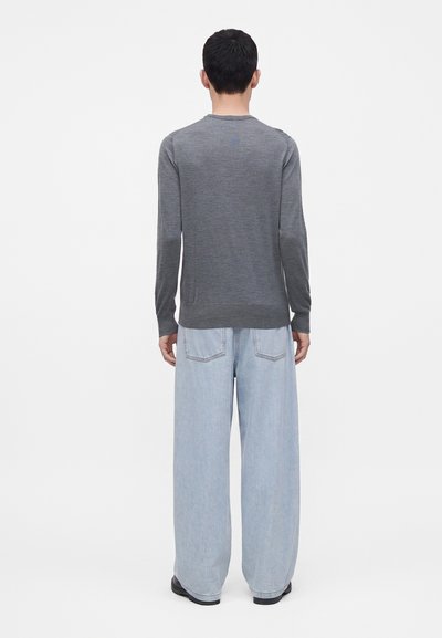 Homme portant une chemise grise à manches longues et un jean large bleu clair, debout de dos sur un fond blanc uni.
