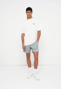 Λευκό oversized t-shirt με μικρή γραφική απεικόνιση, συνδυασμένο με ανοιχτό γκρι σορτς. Λευκά αθλητικά παπούτσια και κάλτσες crew ολοκληρώνουν την εμφάνιση.