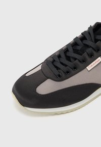 Svart og grå sneakers med en glatt, teksturert overdel, rund tå, svarte snørebånd og en hvit såle med subtilt mønster. Calvin Klein-logo synlig.