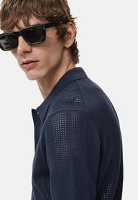 Pulóver azul marino con un patrón texturizado y perforado. Presenta un cuello y mangas cortas. Se lleva con gafas de sol rectangulares negras.