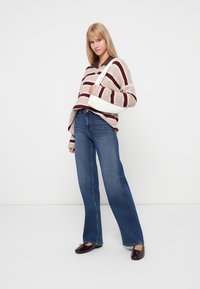 Gebreid sweater met beige en bordeauxrode strepen, v-hals, gecombineerd met blauwe wijde spijkerbroeken. Model houdt een witte handtas vast.