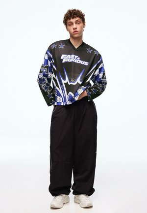 Młody mężczyzna o kręconych włosach, ubrany w czarno-niebieską koszulkę z długim rękawem "Fast & Furious", luźne czarne spodnie i białe trampki, stoi z rękami w kieszeniach.