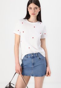 Bijela majica s raštrkanim vezom u obliku crvenih srca, u kombinaciji s plavom denim mini suknjom. Model drži smeđi ručni torbicu.
