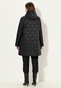 Svart vadderad kappa med huva, med ett diamantmönster, långa ärmar och en avslappnad passform, i kombination med åtsittande svarta byxor och knähöga stövlar.