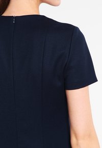 Tmavě modré šaty s krátkými rukávy, hladkým materiálem a zapínáním na zip na zadní straně. Mají přiléhavý střih a jemné švy.