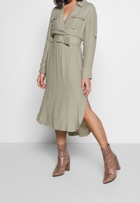 Olivgrön klänning med bälte, långa ärmar och två framfickor, i kombination med ankelstövlar i strukturerat ormskinnsmönster.