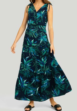 Vrouw draagt een navy blauwe mouwloze maxi-jurk met groene en blauwe tropische bladpatronen en zwarte open teen slip-on sandalen op een lichte achtergrond.