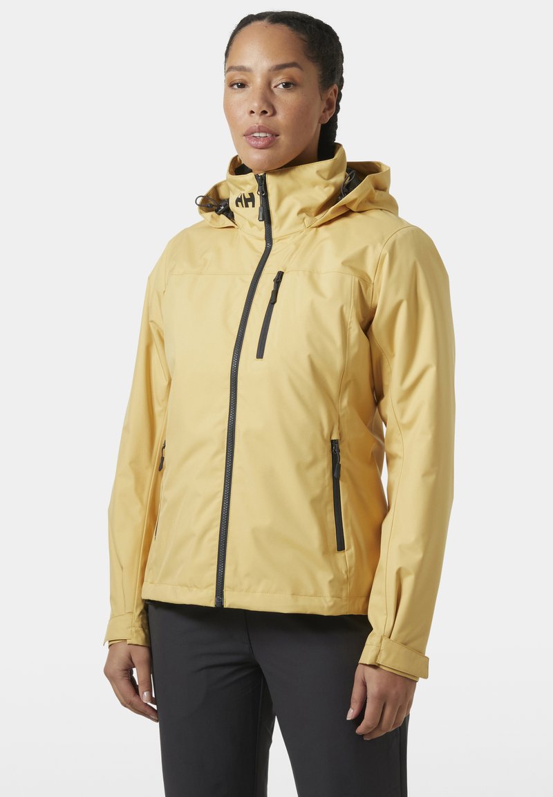 Mujer con chaqueta amarilla con capucha, cremallera negra y bolsillos, de pie frente a un fondo liso.