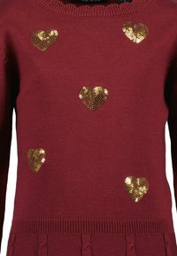 Bordeaux-neule, jossa on kultaiset paljettisydänkoristeet, ribbireunus kaula-aukossa ja helmassa sekä tekstuuri yksityiskohtia alhaalla.