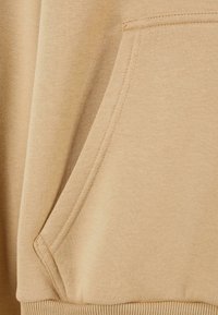 Beige hoodie fabric with a kangaroo pocket, featuring a soft texture and ribbed cuffs. Seam details and smooth finish are visible.