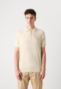 Stickad polo i ljusbeige med korta ärmar, halvmaskipskrage och ribbad fåll. Slät textur med minimalistisk design.