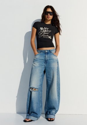 Femeie purtând tricou negru scurt atipic cu grafică, blugi largi oversized rupți, sandale negre și ochelari mari de soare negri, stând în fața unui perete alb.