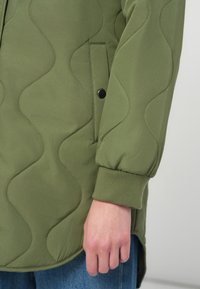 Olīvzaļa quilted jaka ar teksturētu viļņu rakstu, ribotiem piedurkņu galiem un pogu kabatu ar snap-button. Veidota brīvā fitā.