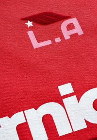 Rød stof med hvid blokbogstaver og en pink "L.A." med en lille hvid stjerne og et maroon bladaccent. Teksturdetaljer synlige.