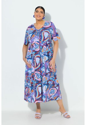 Kobieta w kolorowej sukience midi w paisley wzór z krótkim rękawem i liliowymi sandałami na obcasie, stojąca na jednolitym tle.