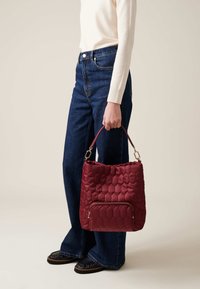 Red quilted handbag with a textured hexagonal pattern, featuring a front zipper pocket and a sturdy shoulder strap. Casual outfit with flared jeans.