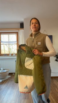 Zaļa un krēmkrāsas jaka ar rāvējslēdzēju, lielu baltu kabatu un trīsstūrveida logotipa detaļu; to tur persona, kas valkā bēša vestu.