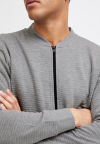 Sweat à zip gris avec texture côtelée, col debout et fermeture éclair noire. Manches longues avec poignets ajustés, sans motifs ni accents visibles.