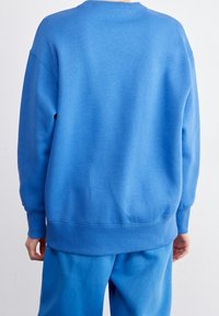 En person som bär en enkel blå sweatshirt och matchande blå byxor, sedd bakifrån mot en neutral bakgrund.
