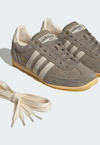 Γκρι σουέντ αθλητικά παπούτσια Adidas Japan με κρεμ ρίγες, λαστιχένιες σόλες και ένα ξεχωριστό ζευγάρι κρεμ κορδόνια δίπλα τους.