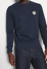 Man som bär en marinblå långärmad sweatshirt med en liten hexagonal logotyp på vänster bröst och mörkblå jeans.
