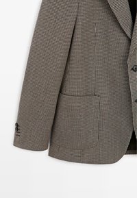 Brązowo-kremowa marynarka w jodełkę z pojedynczą kieszenią i czarnymi guzikami. Posiada klapy z wcięciem i delikatną fakturowaną powierzchnię.