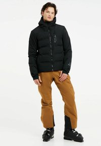 Schwarze, puffy Jacke mit hohem Kragen, Reißverschlusstasche und elastischen Bündchen, kombiniert mit braunen Hosen mit figurnahem Schnitt und schwarzen Akzenten.