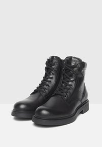 Par de botas de cuero negro con cordones hasta el tobillo, con suelas de goma gruesa y ojales de metal, exhibidas sobre una superficie blanca y un fondo gris.