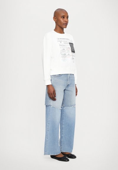 White sweatshirt with graphic prints, paired with light blue denim jeans featuring a patchwork design and frayed edges. Black flat shoes.