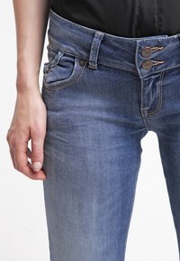 Mörkblå denimjeans har en mid-rise midja med två knappar och en texturerad yta. Designen inkluderar framfickor och kontrasterande sömmar.