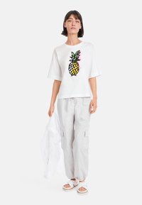 White cotton t-shirt featuring a colorful pineapple graphic, paired with light gray cargo pants and white sandals. Flexible fit.